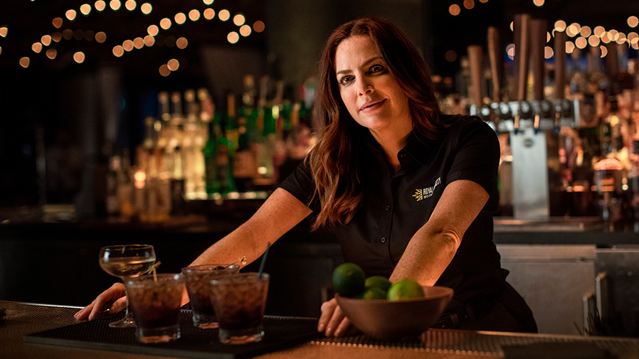 A bartender wearing a personalized Women's UniSport® Micropiqué Polo Shirt provided by UniFirst as part of a UniFirst direct sale program prepares drinks for the guests of the bar.