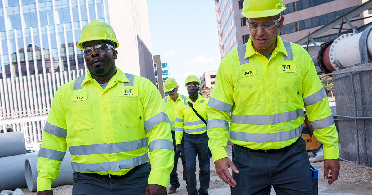Construction workers walking in a city setting while wearing high visibility shirts, hard hats, and safety gear.