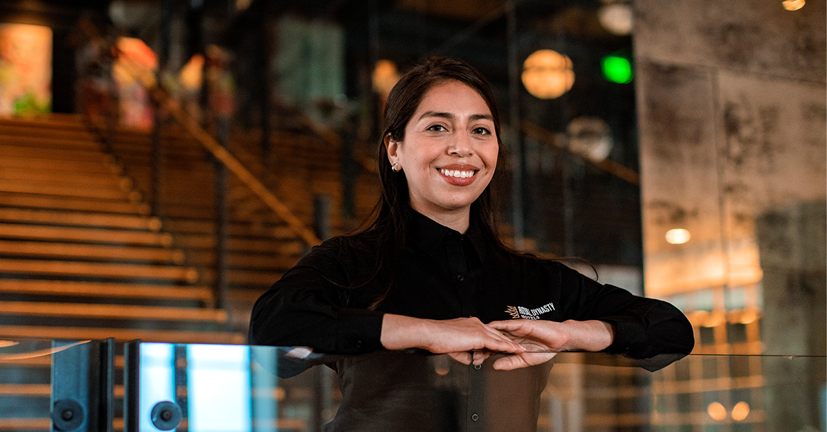 Woman standing at the front desk of a hospitality establishment wearing a black button-down shirt with embroidered company logo.