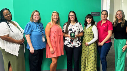 Group photo of seven UniFirst Owensboro Team Partners standing in front of a green wall labeled 'Training Room.' The fourth person from the left holds the Bronze Level Stevie Award for sales and customer service, in recognition of UniFirst Go Careers Program.