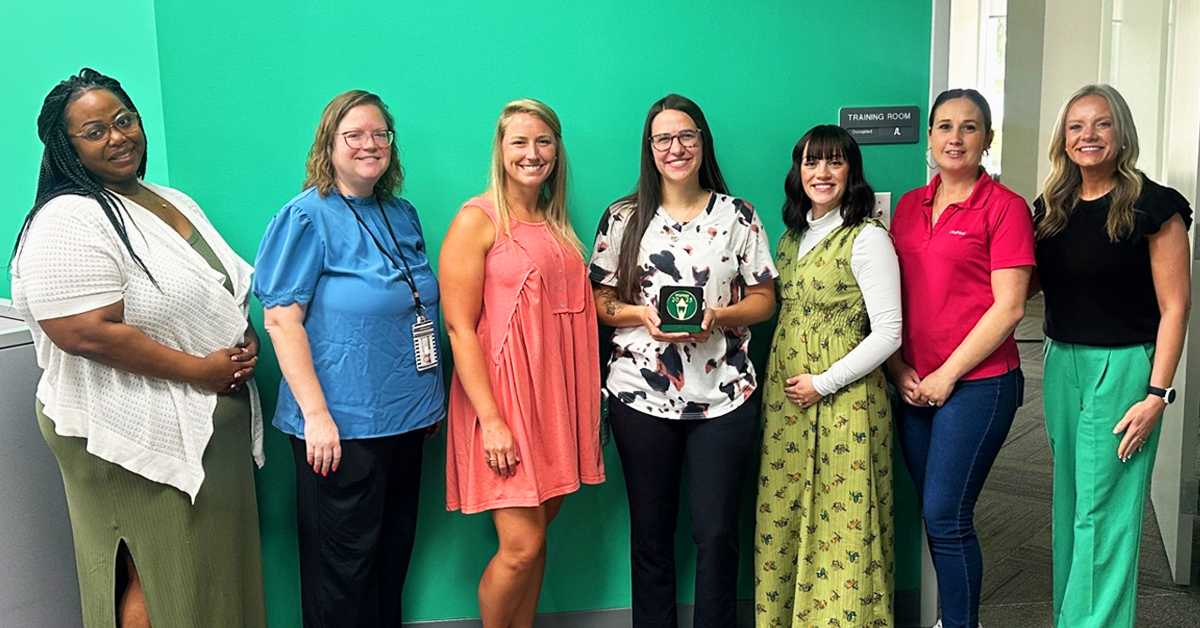 Group photo of seven UniFirst Owensboro Team Partners standing in front of a green wall labeled 'Training Room.' The fourth person from the left holds the Bronze Level Stevie Award for sales and customer service, in recognition of UniFirst Go Careers Program.