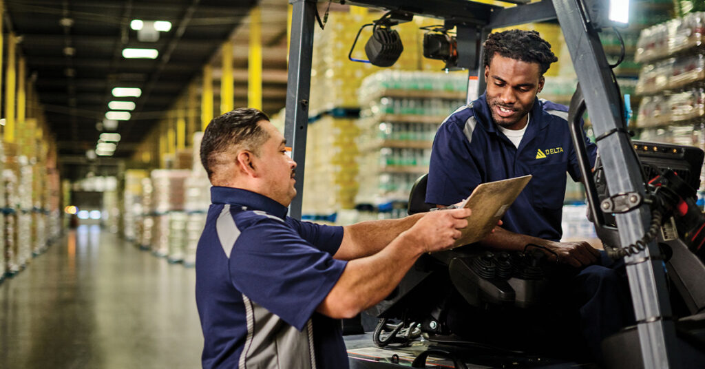 Two men working. One in a forklift and the other showing a clipboard wearing UniFirst uniforms.