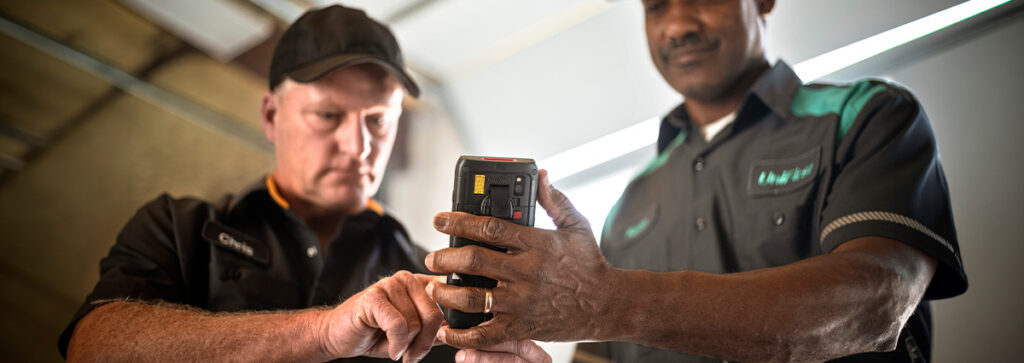 A UniFirst RSR standing with a customer looking over an electronic device showing a UniFirst uniform rental service