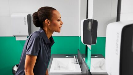 Women stands in commercial restroom pictured with Essential Series Dispensers for soap, paper, and air fresheners.