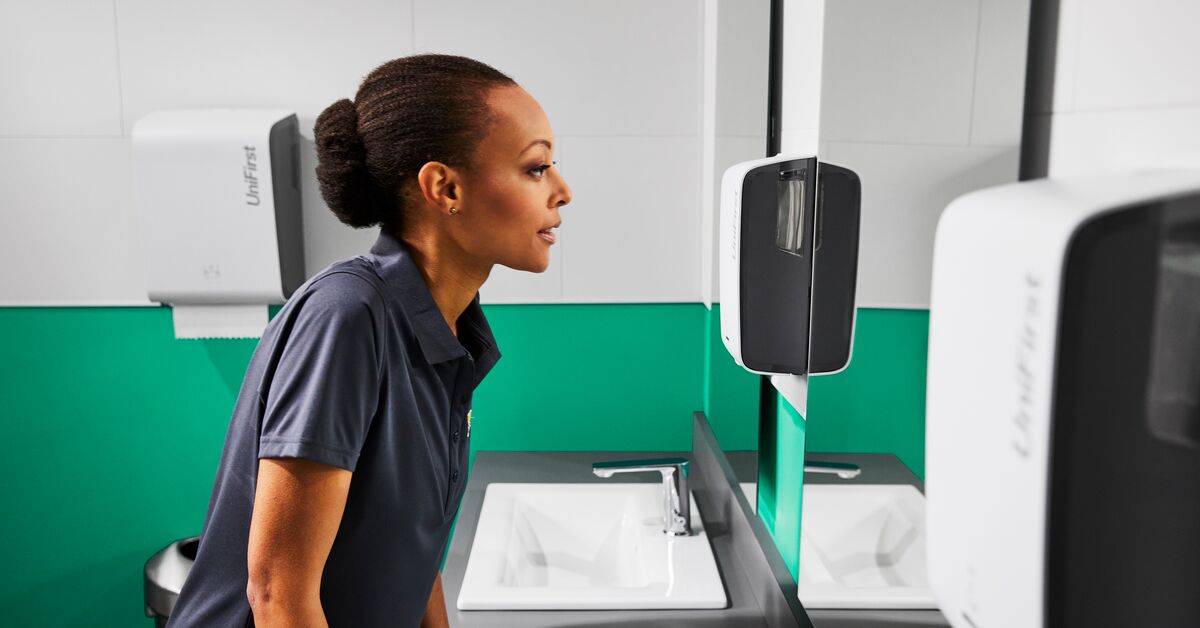 Women stands in commercial restroom pictured with Essential Series Dispensers for soap, paper, and air fresheners.