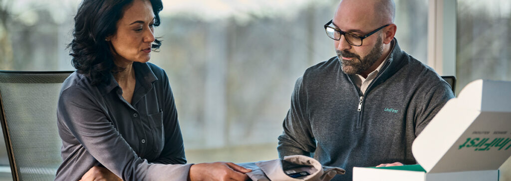 A UniFirst representative and customer sit together at a table looking over a uniform and discussing uniform rental services