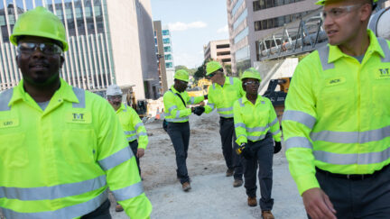 A group of construction workers wearing high visibility clothing and safety apparel walk through a job site, two shaking hands