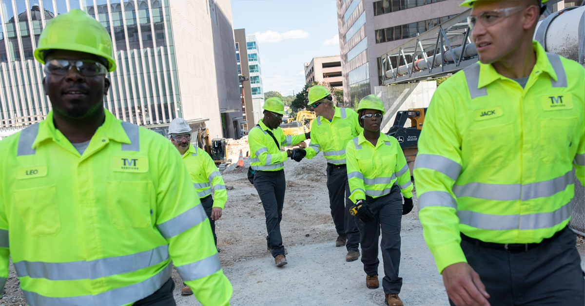 A group of construction workers wearing high visibility clothing and safety apparel walk through a job site, two shaking hands