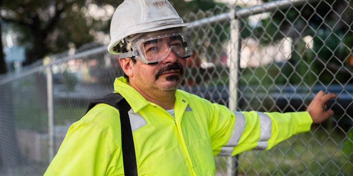 A man wearing ANSI/ISEA 107 clothing stands near a chain link fence