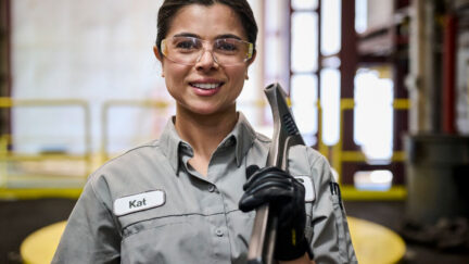 A woman smiling with safety glasses and the tool to get her job done while wearing women's work uniforms
