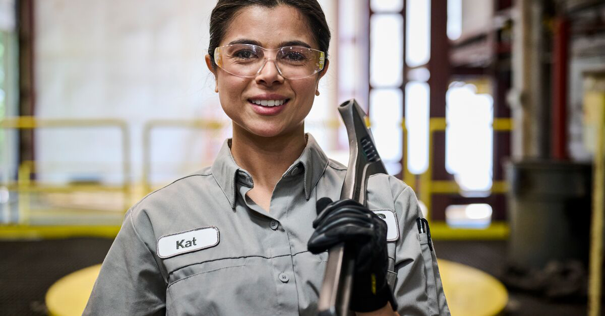 A woman smiling with safety glasses and the tool to get her job done while wearing women's work uniforms